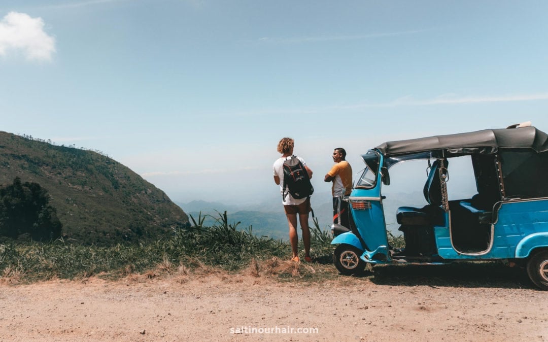 Two people stand near a blue tuk-tuk on a dirt road in Ella Sri Lanka, gazing over a scenic, mountainous landscape under a clear sky.