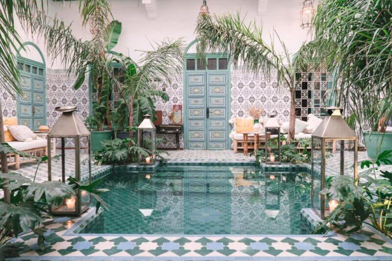 A tiled courtyard with a small pool, surrounded by plants, lanterns, patterned walls, and green doors, featuring seating areas with cushions.