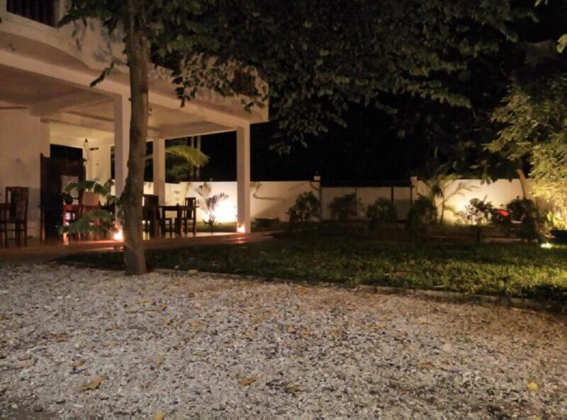 A nighttime view of a patio with tables and chairs, surrounded by a garden and trees in Mirissa, softly illuminated by outdoor lights and accompanied by soothing nature sounds.