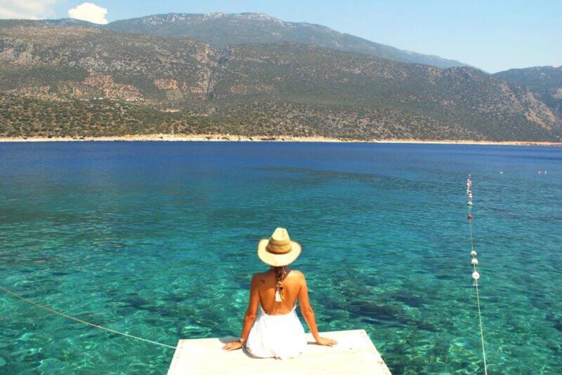 A woman in a sunhat and white dress sits on a dock overlooking the clear turquoise water of Kas-Barbarossa, with mountains and a blue sky in the background.