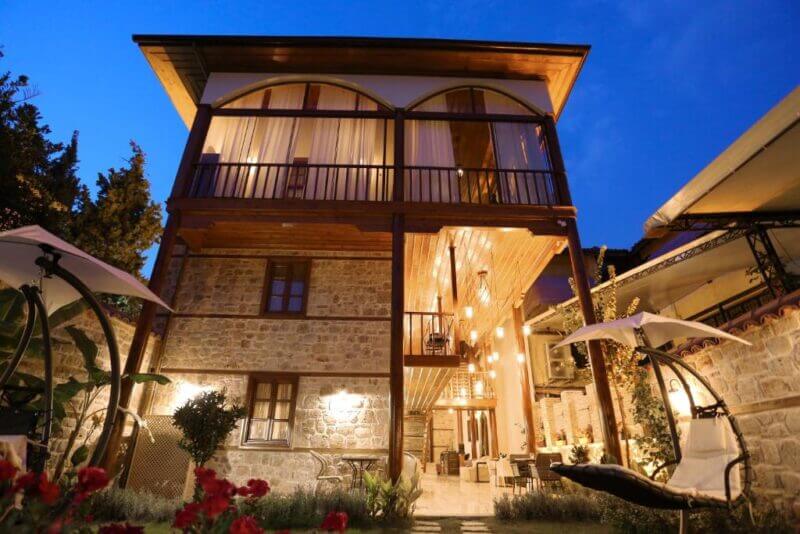 Three-story stone and wood char-me-antalya house with large windows and balconies, warmly lit at dusk, featuring outdoor seating and lush garden plants in the foreground.