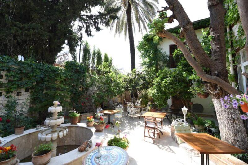 A sunny garden courtyard at Sibel Pension in Antalya, featuring a central fountain, potted plants, outdoor tables and chairs, all surrounded by lush trees and greenery.