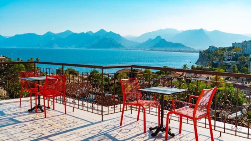 Rooftop terrace at Patron Hotel with red chairs and tables overlooks a blue sea, distant mountains, and a coastal town under a clear sky.
