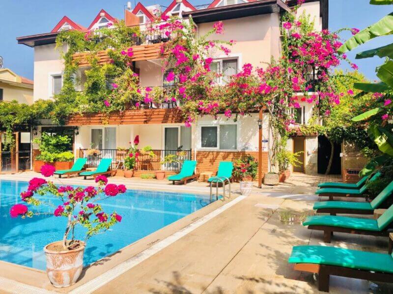 A sunlit hotel pool area at Ten Apart Fethiye features turquoise lounge chairs, potted plants, and a building covered in vibrant pink bougainvillea flowers.