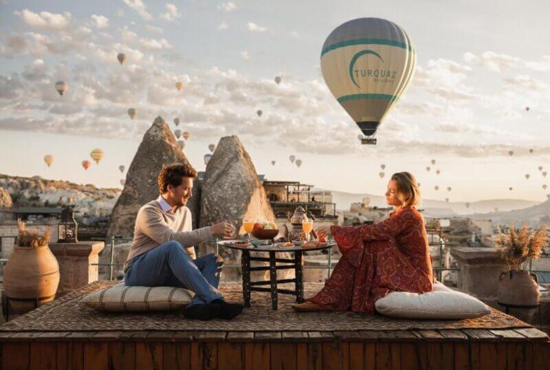 A man and woman sit on cushions at a low outdoor table at Sultan Cave Suites, sharing a meal as hot air balloons rise over the rocky landscape at sunset.