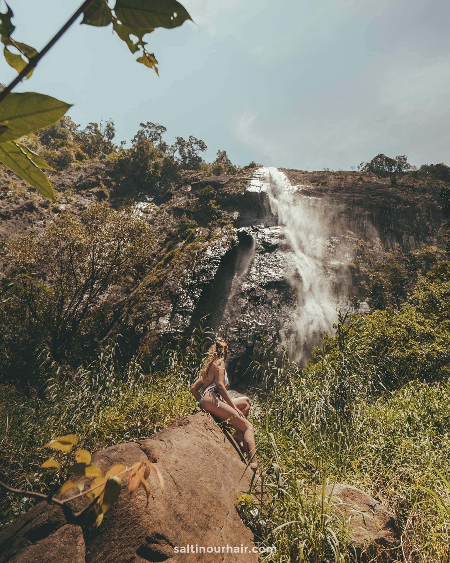 A woman in swimwear sits on a rock before Diyaluma Falls, Sri Lanka, surrounded by lush greenery and a rocky cliff beneath a partly cloudy sky.