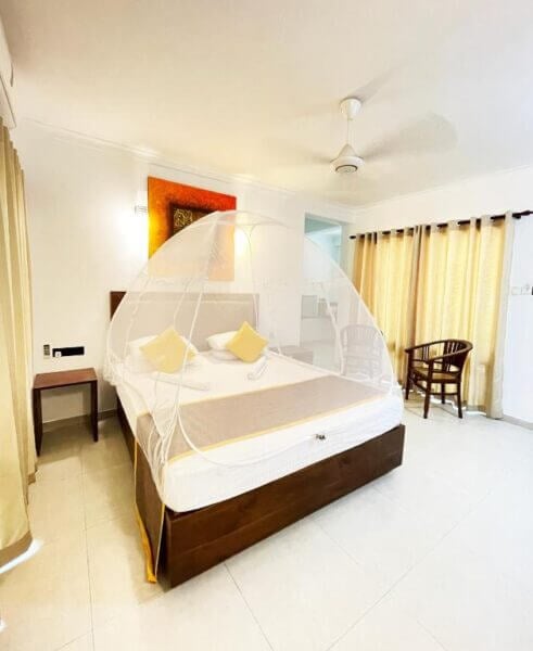 A bright hotel room in Dambulla with a double bed covered by a white mosquito net tent, two chairs, a ceiling fan, and large windows with beige curtains overlooks a stunning rock arch formation.