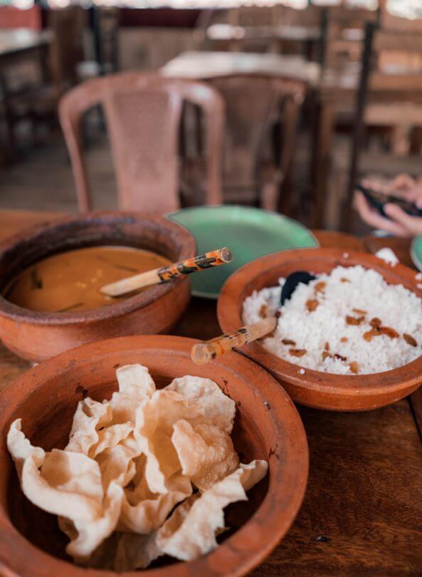 Three clay bowls on a wooden table feature curry, white rice with raisins, and crispy papadum&mdash;a taste of Trincomalee, Sri Lanka. Two green plates and wooden chairs are visible in the background.