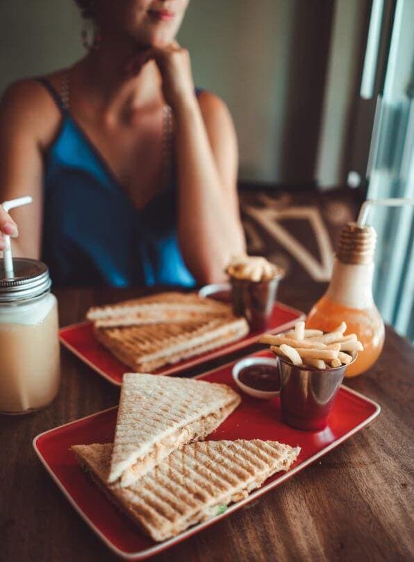 A person in a blue top sits at a table in Trincomalee, Sri Lanka, with two plates of grilled sandwiches, fries, ketchup, and drinks in glass jars with straws.