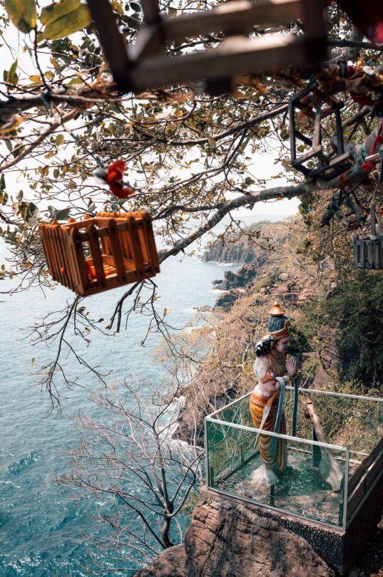 A statue of a woman in traditional attire stands on a glass platform overlooking the sea near Koneswaram Temple Trincomalee, with bird cages and a wooden swing hanging from tree branches above.
