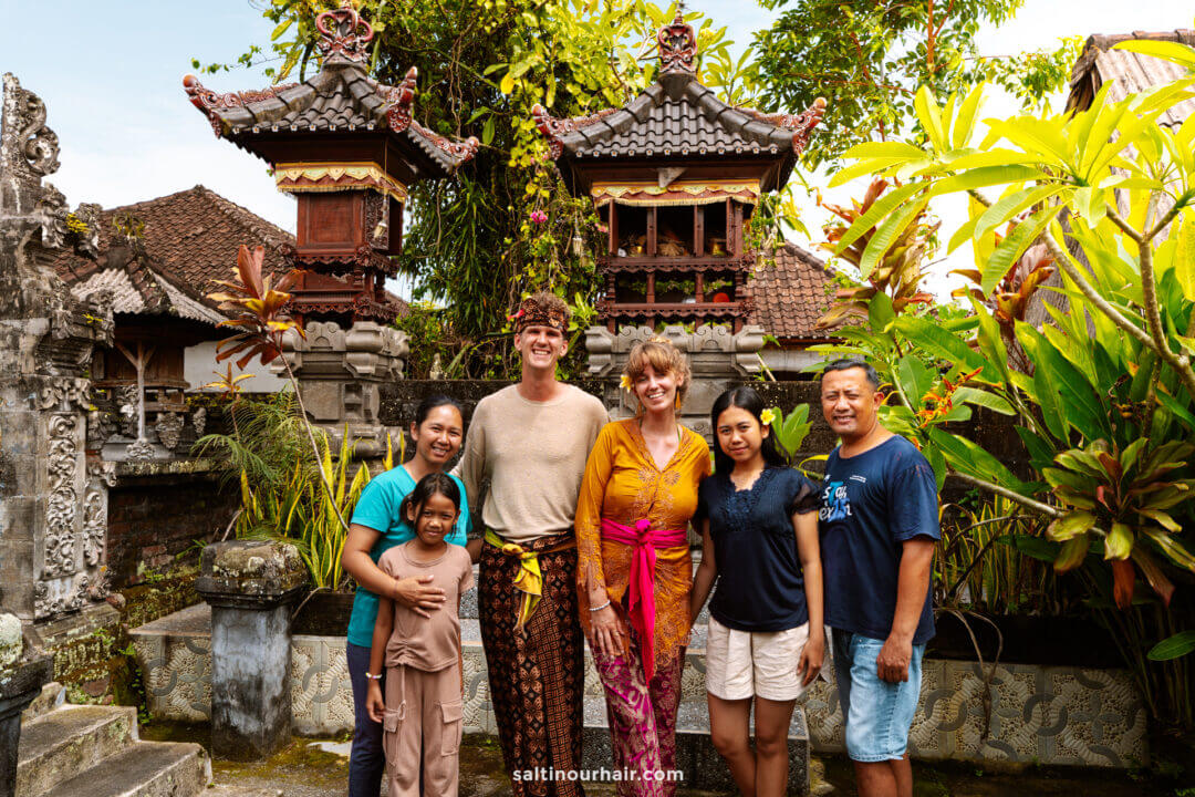 Six people stand together smiling in front of traditional Balinese architecture at a charming homestay Bali, surrounded by lush greenery and intricate stone carvings.