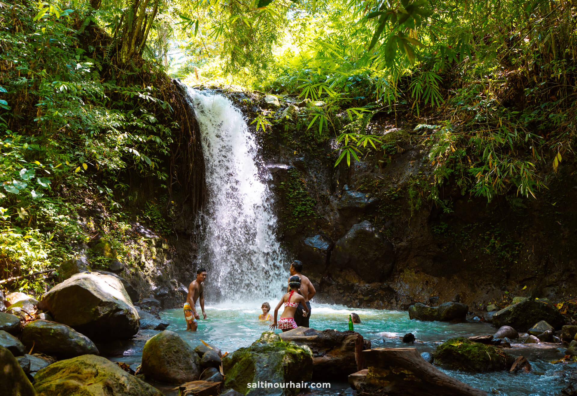 A small group of people swim and stand in a turquoise pool at the base of a waterfall, surrounded by lush green vegetation, experiencing the natural beauty near a homestay Bali offers.