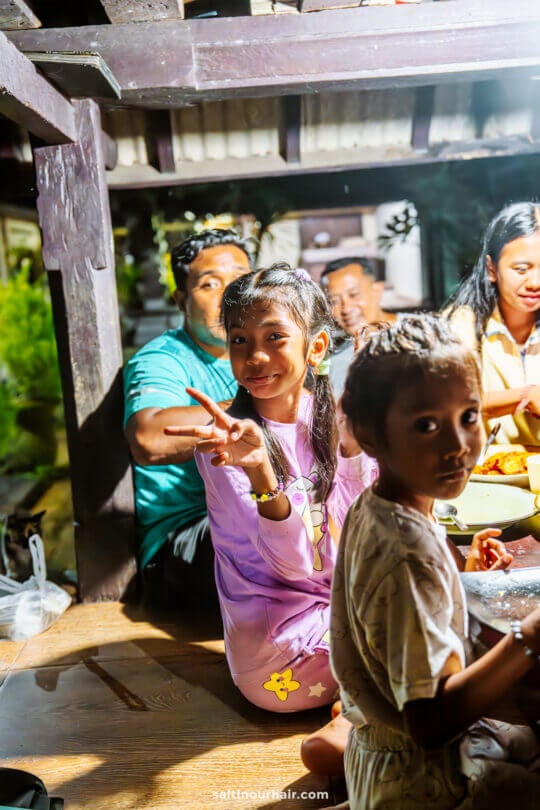 A group of people sit together indoors at a homestay in Bali, with a young girl in the foreground smiling and making a peace sign, while another child looks toward the camera. Food is visible on the table.