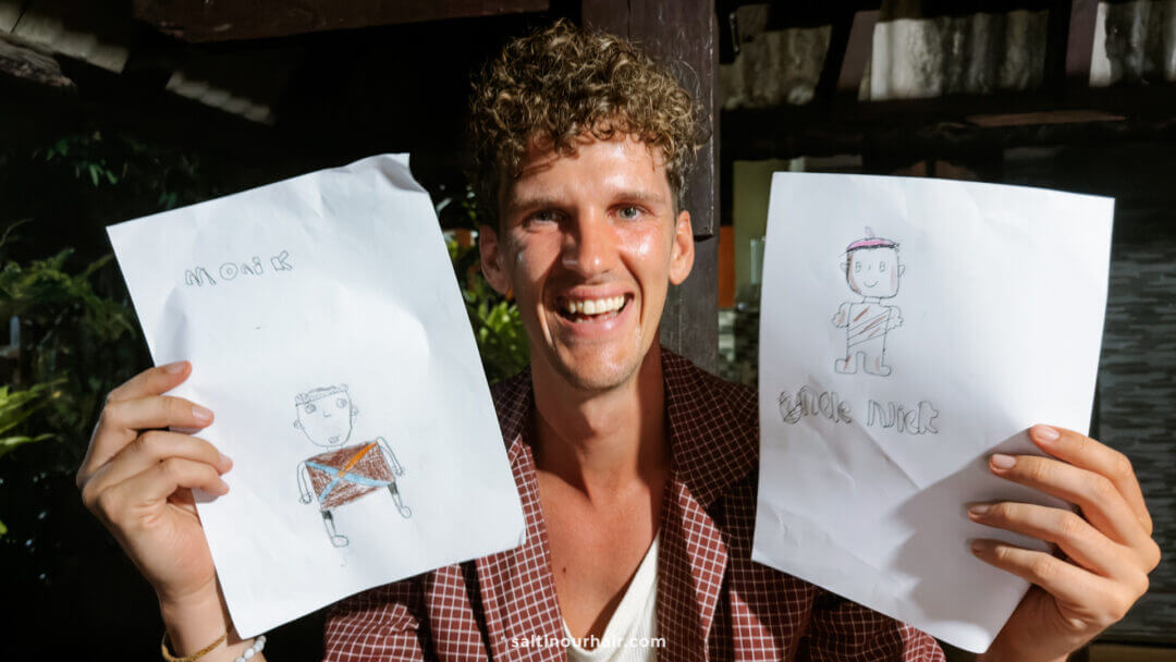 A person smiling and holding up two hand-drawn pictures of people, each labeled with text above the drawings, captures the welcoming spirit often found at a homestay Bali experience.