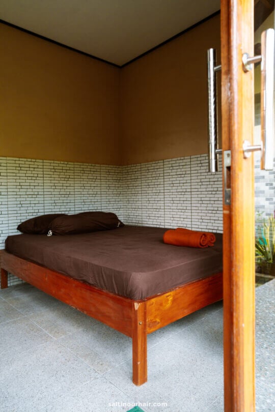 A simple wooden bed with brown sheets and a rolled orange towel in a minimalist homestay Bali room, featuring white brick-patterned walls and an open wooden door.