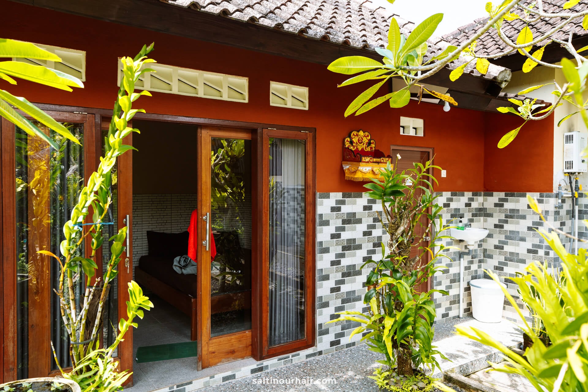 A small homestay Bali bungalow with an open glass door, tiled exterior walls, lush plants in front, and a visible bed and sink area.