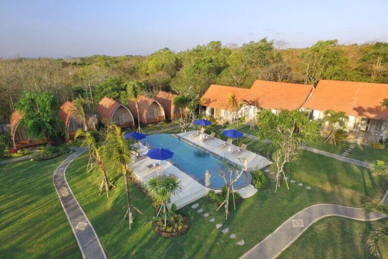 Aerial view of Bakal Sari Uluwatu resort with bungalow-style cabins, a central swimming pool shaded by blue umbrellas, lush greenery, and inviting stone pathways.
