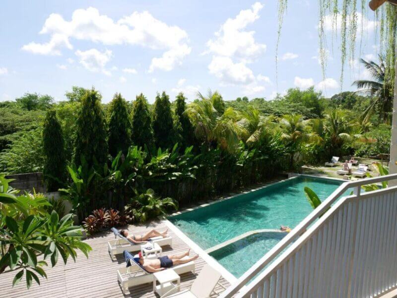 Two people sunbathe on lounge chairs beside a rectangular outdoor pool at an inn near Padang Padang, surrounded by tropical plants on a sunny Uluwatu day.