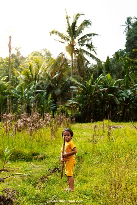 A young child in a yellow outfit stands in a grassy field holding a stick, with palm trees and lush greenery from a homestay Bali setting in the background.