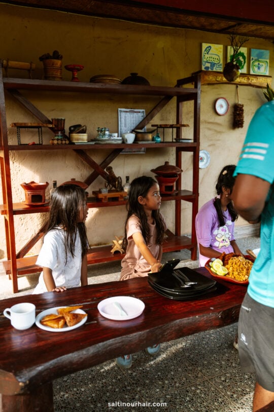 Three children sit at a wooden table with plates of food, while an adult stands nearby. Shelves with various items are visible in the background, capturing a cozy homestay Bali atmosphere.
