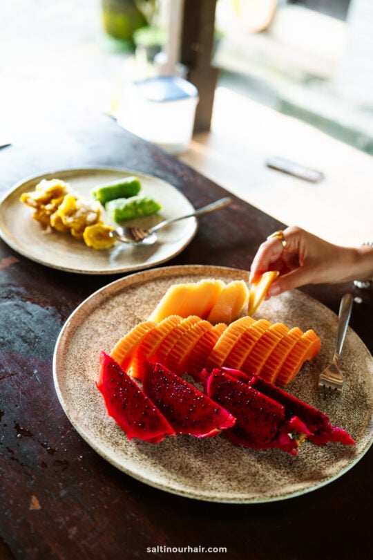 A hand with a fork reaches for sliced cantaloupe and dragon fruit on a plate at a cozy homestay Bali breakfast; another plate with assorted pastries is in the background.