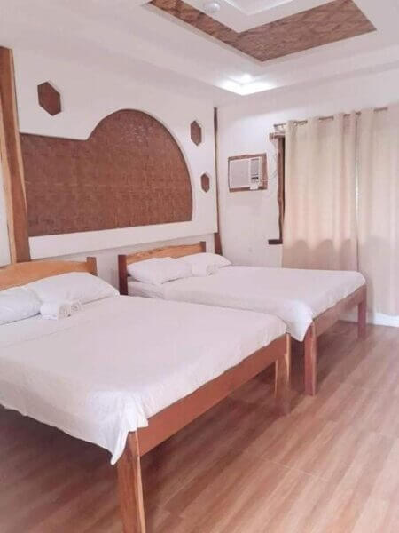 Two wooden beds with white linens and rolled towels in a simple room featuring wooden flooring, wall decor, an air conditioner, beige curtains covering a window—bringing a touch of Siquijor sand’s natural charm indoors.
