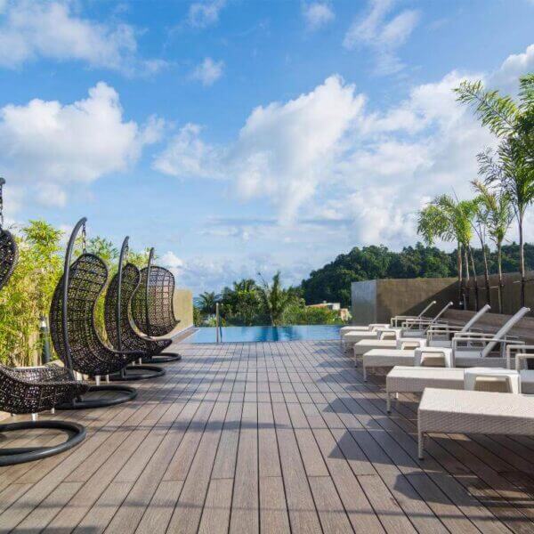 Rooftop pool area at Cuna Resort with wicker egg chairs, white lounge chairs, wooden deck, palm plants, and a view of greenery under a blue sky with clouds.