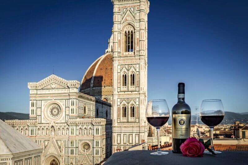 A bottle of red wine, two filled glasses, and a pink rose rest on a ledge at la-terrazza-duomo, overlooking Florence’s Cathedral of Santa Maria del Fiore and bell tower beneath a clear sky.