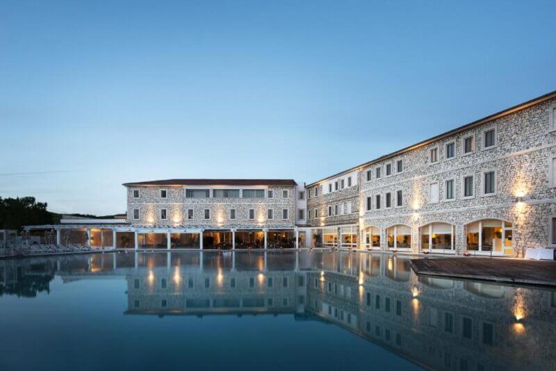 A large stone building with multiple windows is reflected in a calm outdoor pool at dusk, with lights illuminating the exterior—evoking the serene ambiance of a wellness retreat like Saturnia Spa.