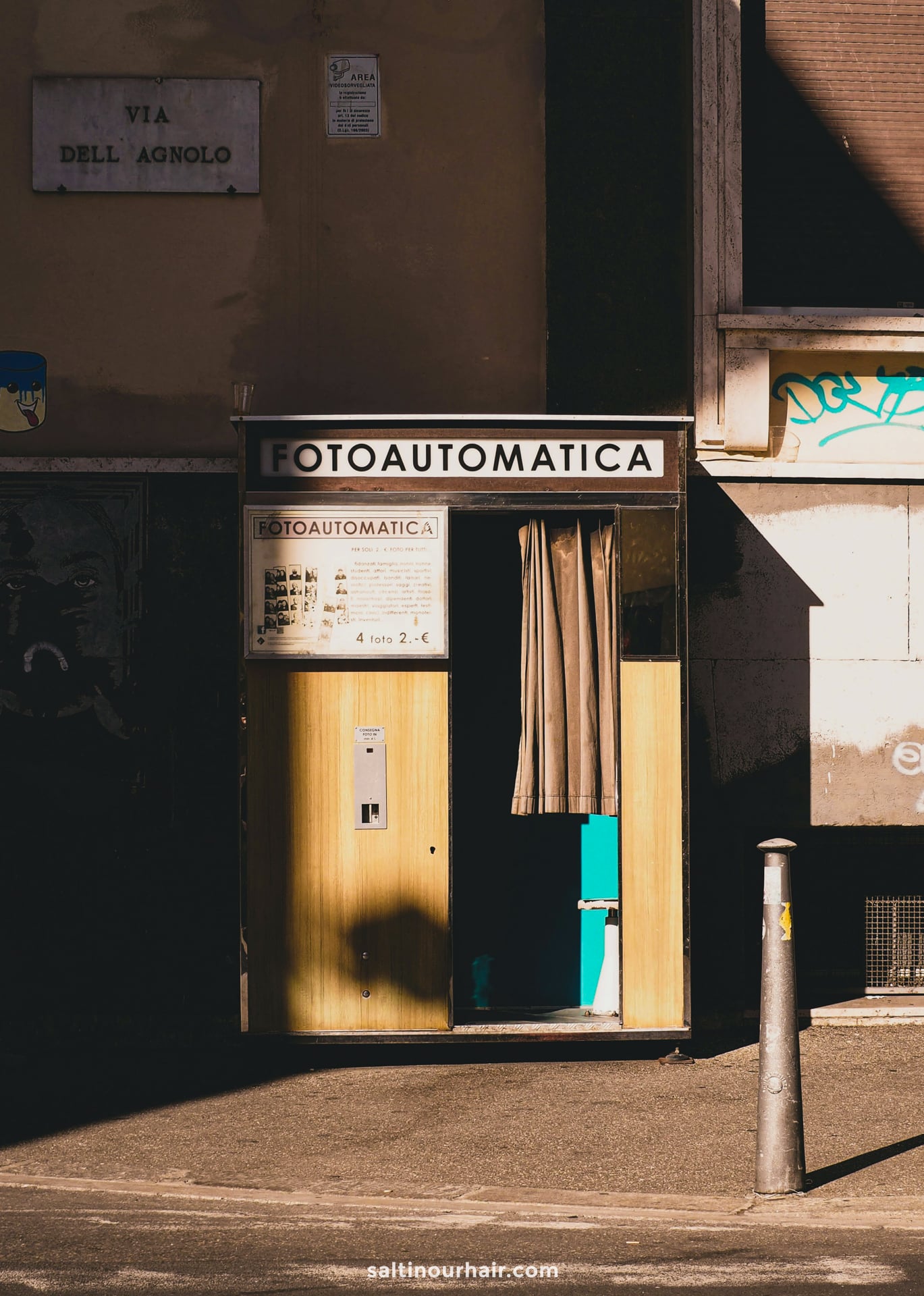 A vintage Fotoautomatica photo booth stands on a sunlit Florence street corner, its curtain partially open and signage displayed above and beside the booth.