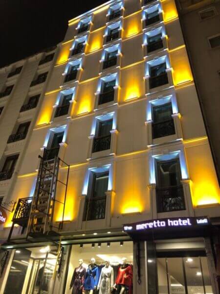 A multi-story hotel building in Istanbul with illuminated yellow and white exterior lights at night and mannequins displayed in the ground-floor windows. The sign reads Meretto Hotel.