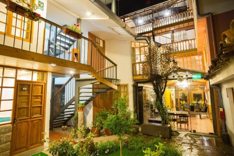Well-lit, multi-level courtyard of a building with wooden doors, metal stairs, indoor plants, and seating area visible through large windows at night.