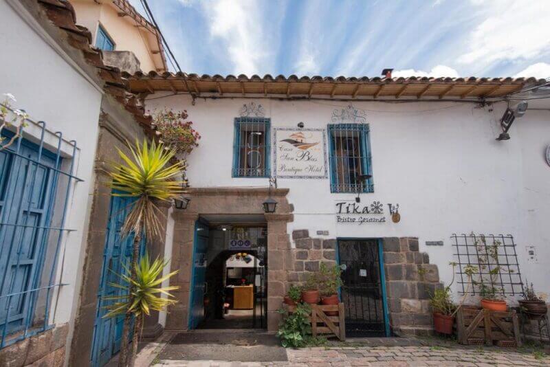 A white-walled building with blue-trimmed windows and doors in San Blas, Cusco, features a boutique hotel sign and potted plants along the stone-paved entrance under a partly cloudy sky.