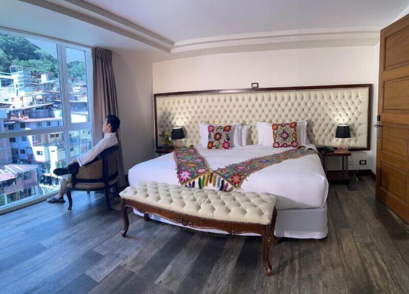 A man sits by the window in a hotel room with a large bed, colorful pillows and runners, tufted headboard, bench, and two bedside tables with lamps.