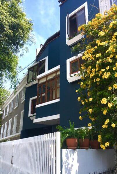 Modern blue and white building with open windows, potted plants, and a wall covered in yellow flowers next to a white fence under a clear sky.