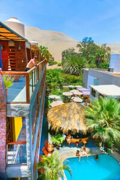 A person stands on a hotel balcony overlooking a courtyard with palm trees, sun loungers, a thatched-roof hut, and a swimming pool where people are relaxing, evoking the laid-back vibe of Huacachina while enjoying a fresh banana smoothie.