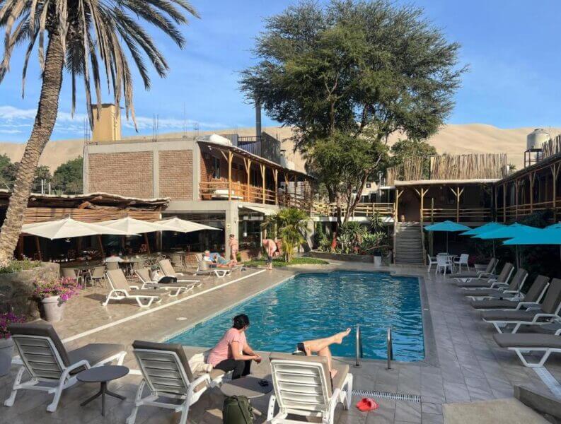 Outdoor hotel pool area at El Huacachinero with lounge chairs, umbrellas, and guests relaxing; surrounded by buildings and palm trees in huacachina, with sand dunes visible in the background.