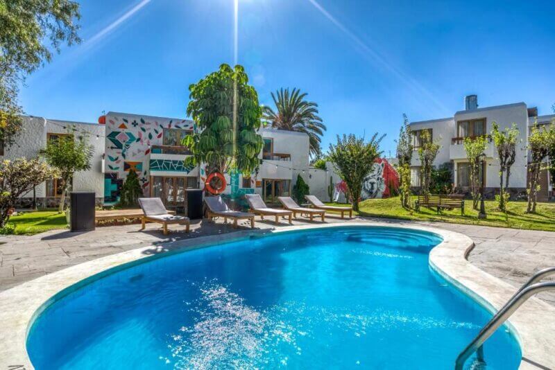 A bright outdoor pool with lounge chairs beside it at Selina Arequipa, surrounded by grass, trees, and modern white buildings under a clear blue sky.