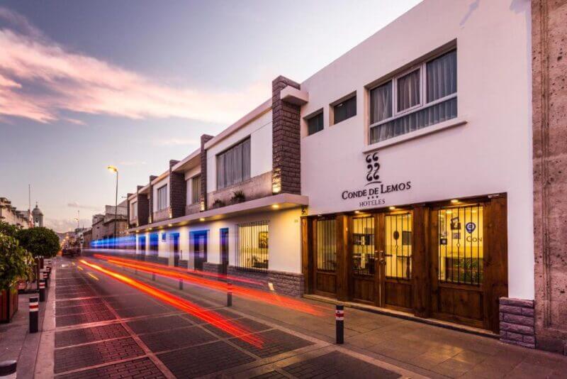 Modern white hotel building with wooden doors and stone accents at dusk in Arequipa, with light trails from passing vehicles visible on the street—offering a stylish stay near Lemos.