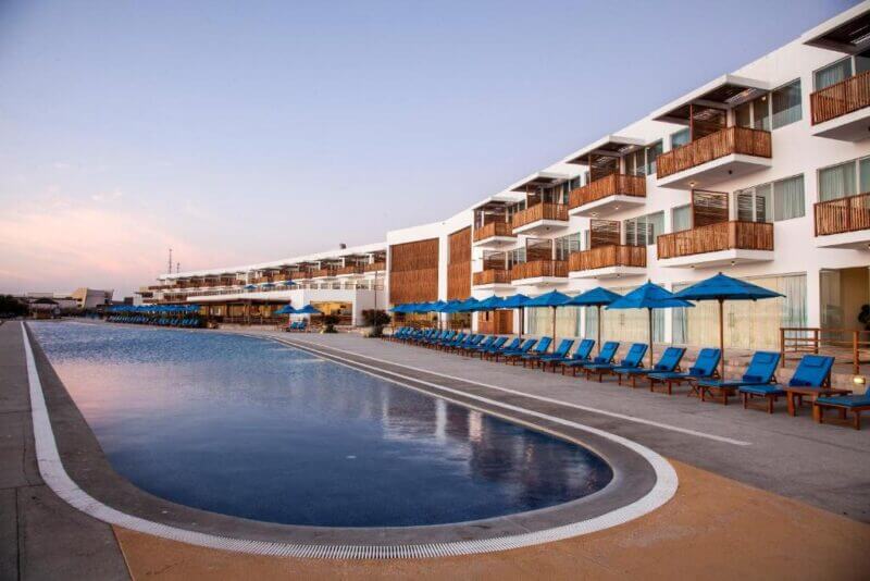 Outdoor hotel pool at San Agustin Paracas with rows of blue lounge chairs and umbrellas beside a modern multi-story white building with balconies at sunset.