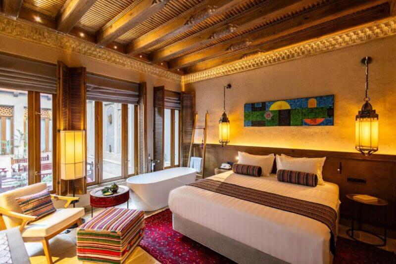 A warmly lit hotel room at Kukaldosh in Bukhara features a king bed, modern bathtub, wooden ceiling beams, large windows, colorful decor, and traditional furnishings.
