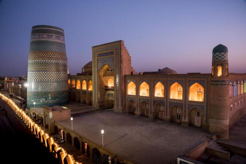 Historic Islamic architecture at dusk features the illuminated Kalta Minor Minaret and surrounding buildings in Khiva, Uzbekistan, near the renowned Orient-Star hotel, with arched windows and intricate tile work.