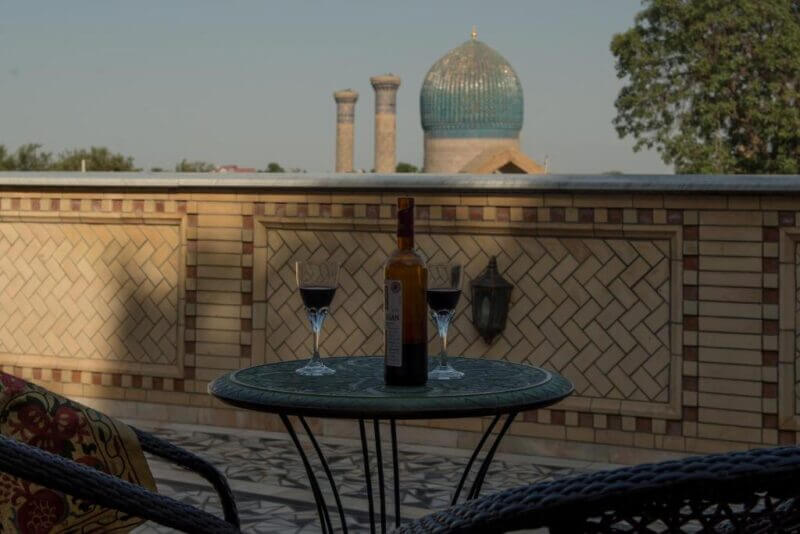 A round table with a wine bottle and two glasses on a balcony at ARGAMAK Samarkand, overlooking a domed building and twin towers in the background.