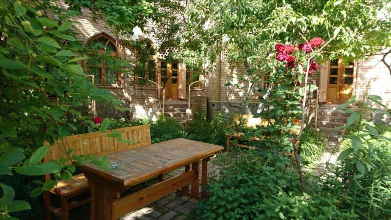 Wooden table and benches sit on a stone patio surrounded by lush green plants and trees at Antica BB Samarkand, with a brick building and arched windows in the background.