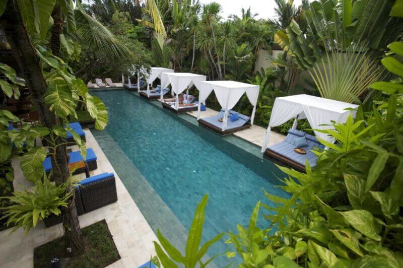 A rectangular outdoor pool at Elysian Seminyak is surrounded by lush greenery and lined with canopy daybeds featuring blue cushions on one side.