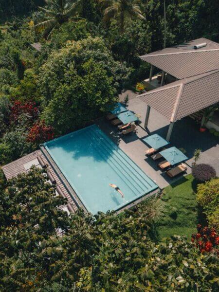 A person floats in a rectangular pool at Aarunya Resort, beside a modern house surrounded by lush greenery and outdoor seating with umbrellas in Kandy.