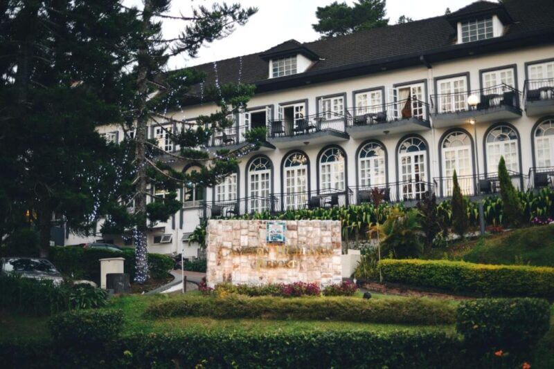 A large white building with black trim and multiple arched windows stands behind a stone sign and landscaped greenery, offering an inviting glimpse of the elegant Cameron Highlands Resort.