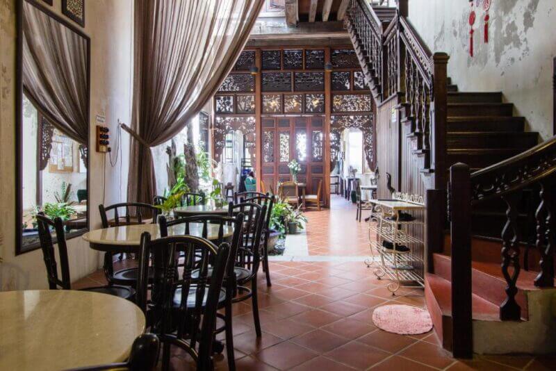 Interior of a traditional cafe at Carnarvon House in Georgetown, featuring wooden furniture, tiled floor, decorative screens, large windows, potted plants, and a wooden staircase on the right.