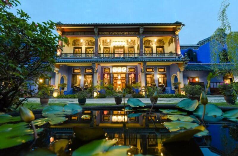 A two-story colonial-style building with ornate details, hotel-cheong-fatt-tze is warmly lit at dusk, its glow reflected in a pond with lily pads in the foreground.