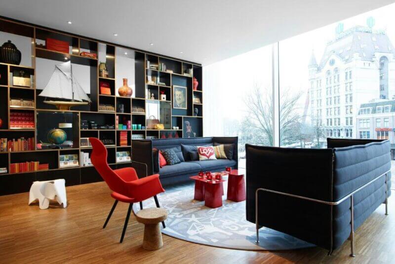 Modern living room with large windows, black sofas, red accents, and bookshelves filled with decor—enjoy a stunning view of a historic building outside in this stylish hotel Rotterdam setting.